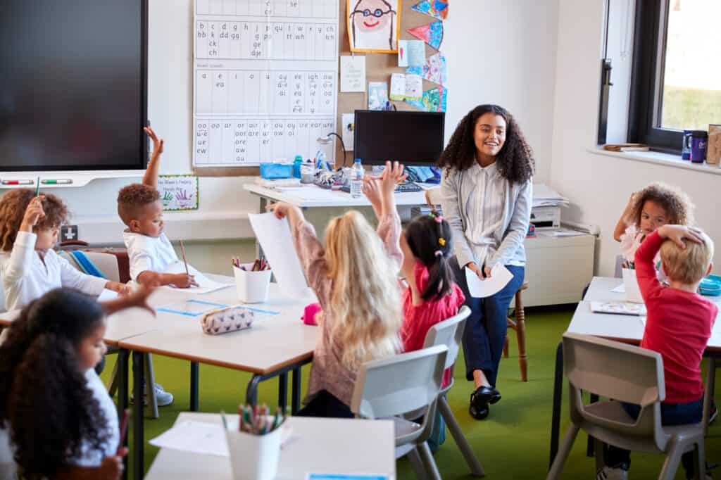 School Based Programs 3 Young Female Infant School Teacher Sitting On A Chair Facing School Kids In A Classroom